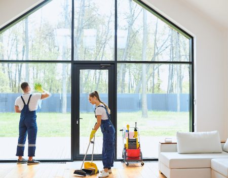 Young cleaners in blue uniform mopping wooden floor, cleaning big panoramic window, preparing country house for selling. Successful cleaning service concept