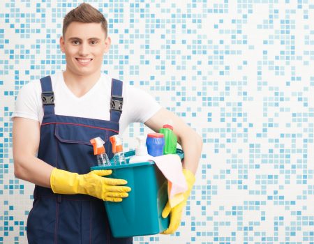 Half-length portrait of young dark-haired smiling janitor wearing white T-shirt blue overalls and yellow rubber gloves holding blue pail with cleaners standing in the bathroom